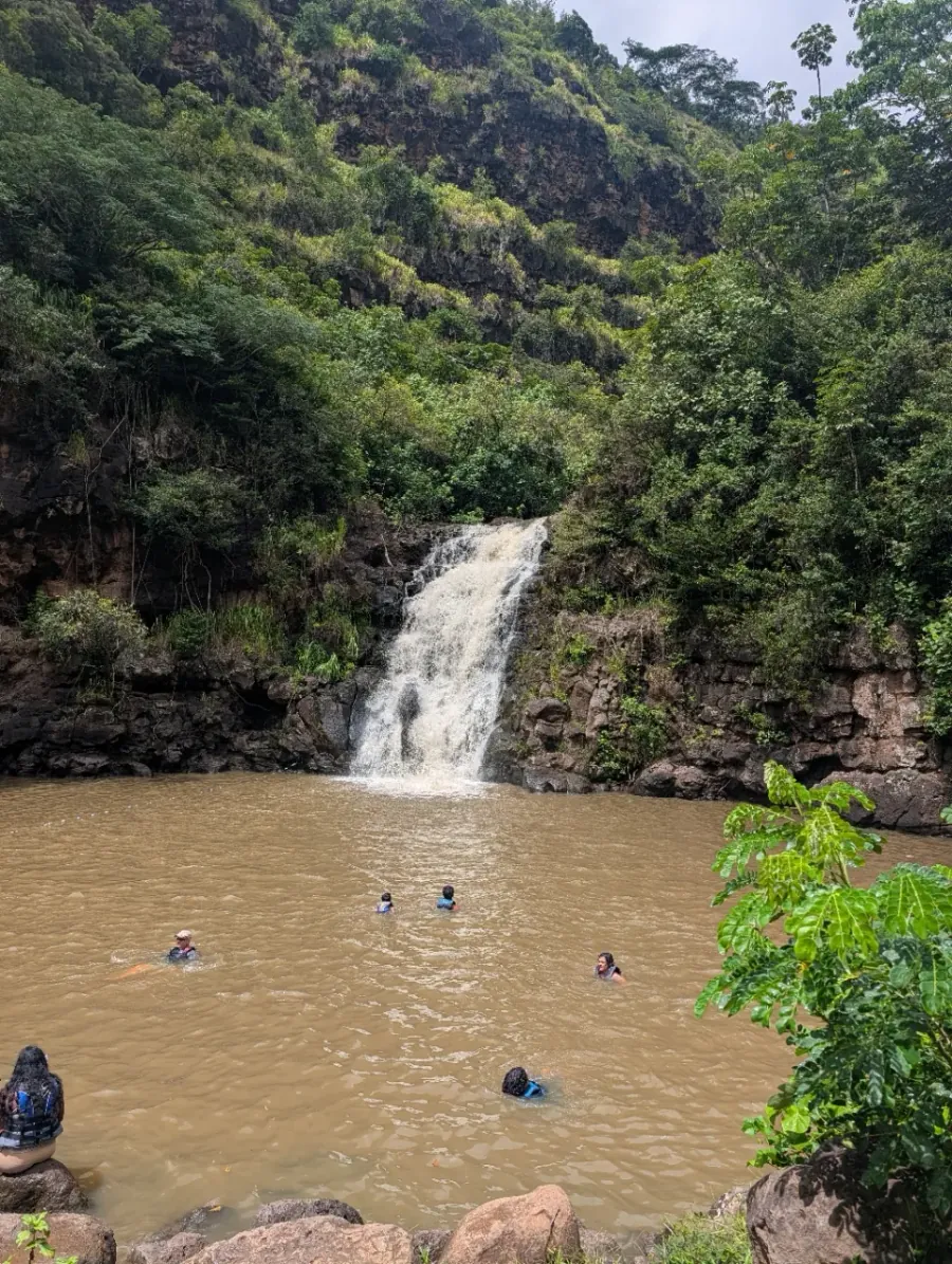 A waterfall on Oahu's North Shore falling into a wide brown swimming pool, with swimmers in life vests and steep green valley walls behind it.